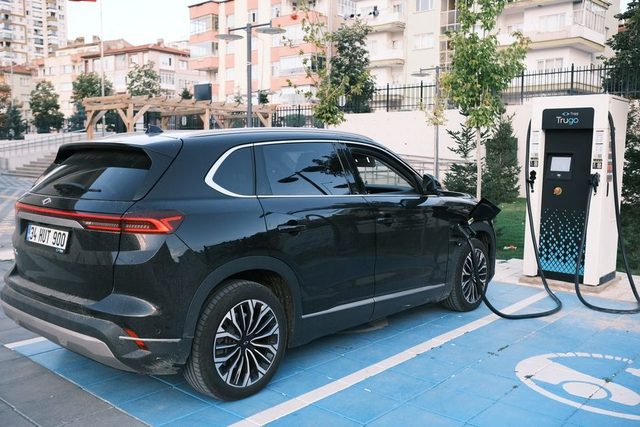 Une voiture électrique d’occasion gris métallisé en 3/4 avant affiche son prix devant une borne en concession